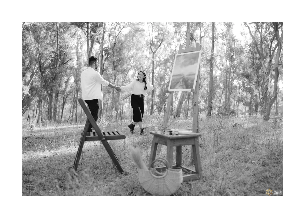 A black-and-white photo of a couple at an easel in a field of trees