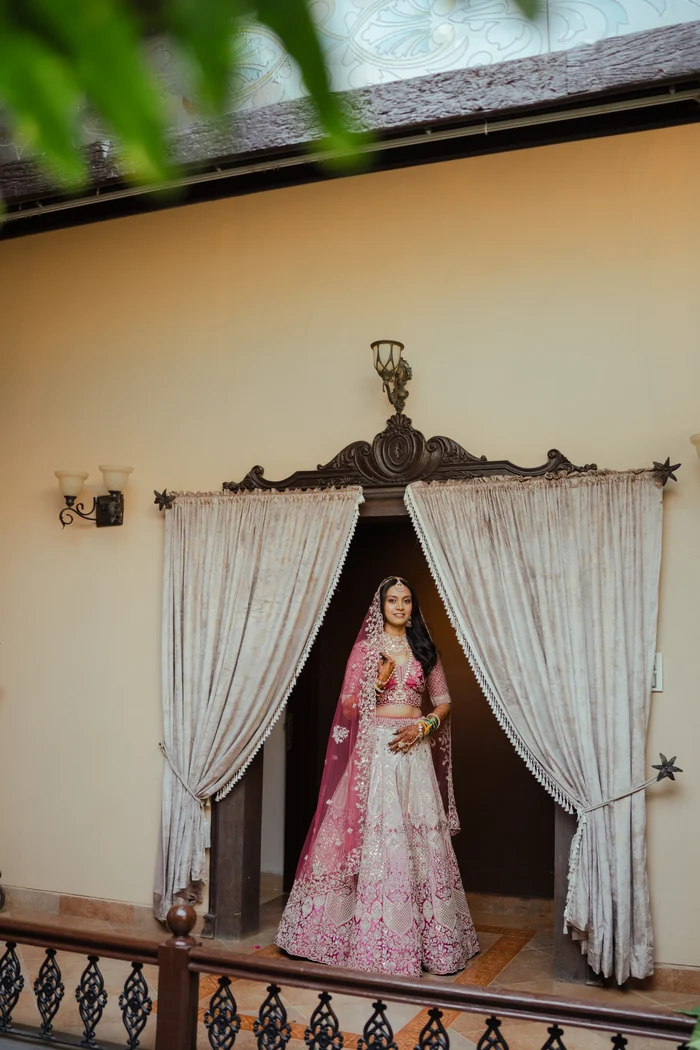 A bride in pink framed by curtains on a balcony